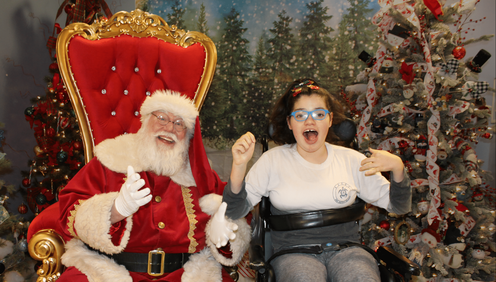 Santa Claus sits in a red throne next to an excited girl in a wheelchair
